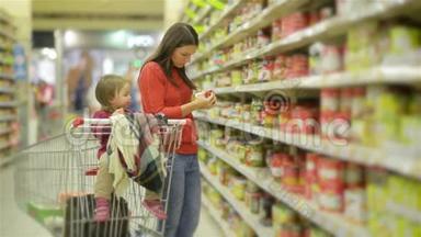 年轻的母亲和孩子沿着批发货架走着，在商店的手推车里取货，女人站在超市附近。